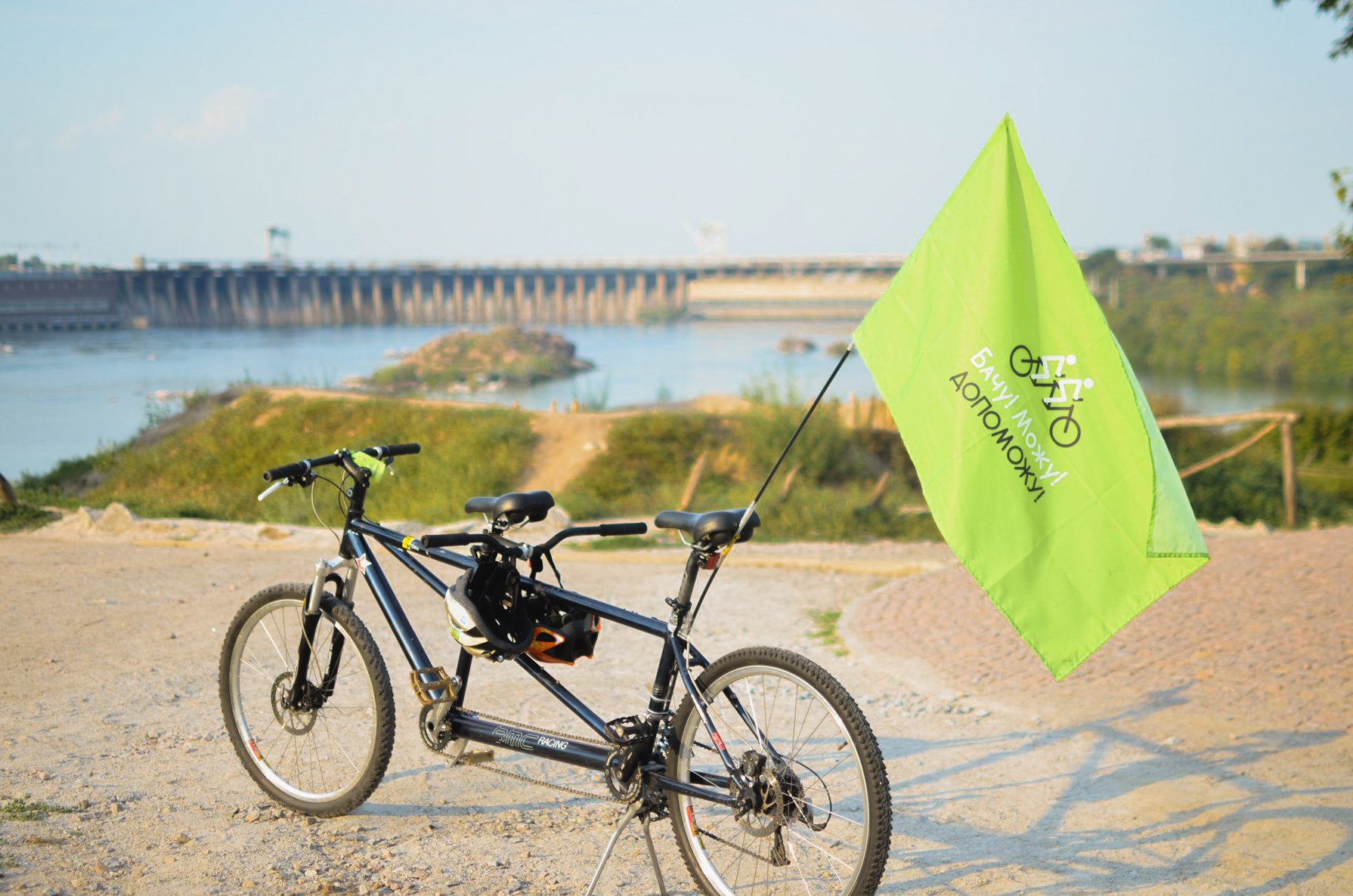  A black tandem bicycle having a green banner with the writing “I see! I can! I will!” in Ukrainian is standing on a path. In the background there is a river and a barrage.