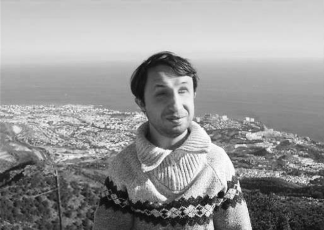 The picture is in black and white. A man is standing on a hill. He is wearing a pullover. His hair is dark. In his back there is a city and a coast. The sun is shining.